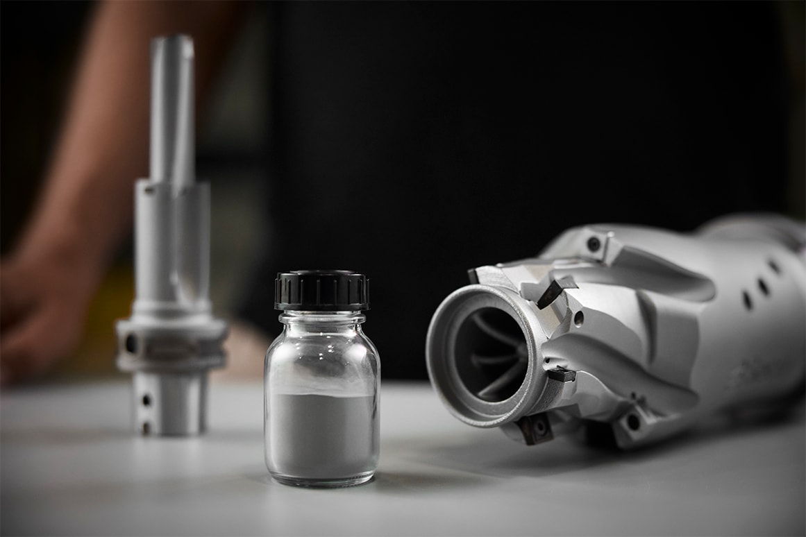 Glass jar with black lid, containing Osprey® MAR 55 metal powder. On each side of the jar are printed parts, courtesy of Seco.