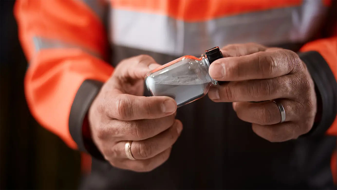 A close-up of a set of hands holding a small, glass jar with black lid, containing grey metal powder.
